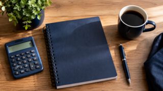 木目の机に置かれた紺色のノート、電卓、コーヒー、観葉植物。家計とお金の計画のイメージ